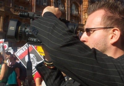 A person with dark hair and glasses, holding a video camera, documenting an anti-war demonstration, holding a video camera surrounded by placards.