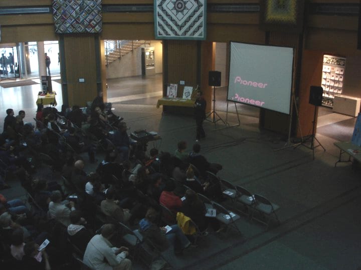 A person in front of a screen, holding a microphone and speaking to a seated audience in an atrium.