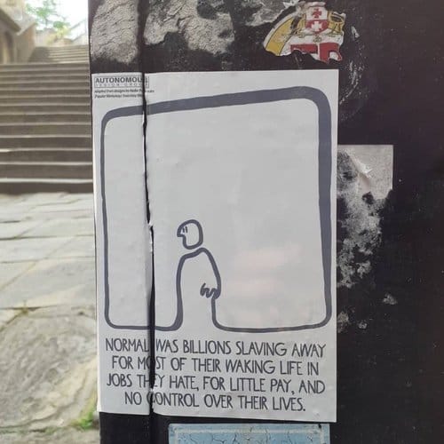 A street with a sticker in foreground with an outline sketch of a person and text "normal was billions slaving away for most of their waking life in jobs they hate, for little pay, and no control over their lives."