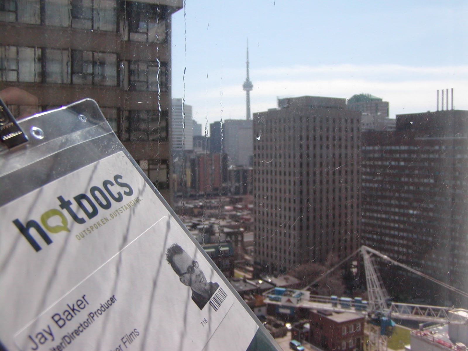 A Hot Docs access pass featuring Jay Baker's name and photo, left in a hotel window with raindrops on it, the CN Tower in the distance