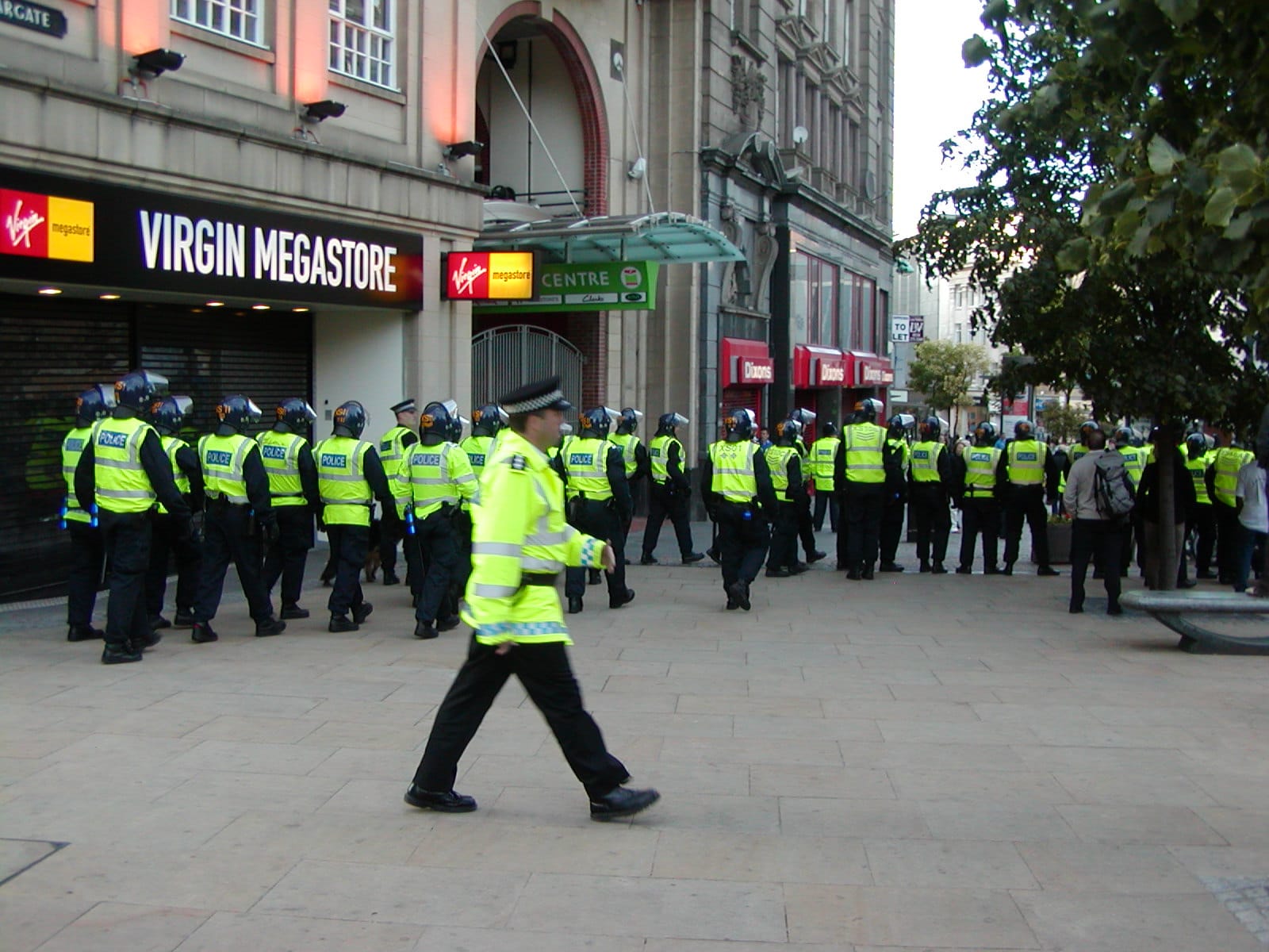 Dozens of armoured cops with one police officer striding behind them in the street.