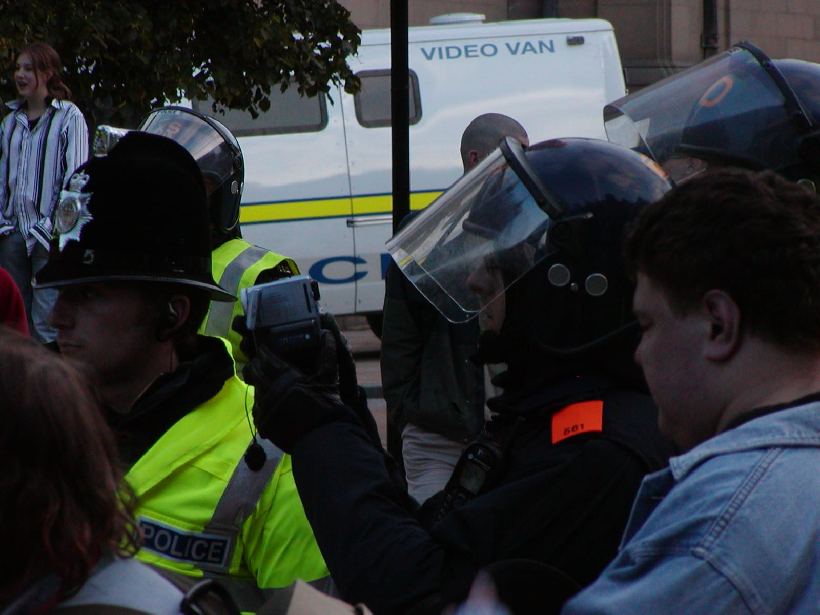 A police surveillance van with armoured cops in front of it, one of whom is filming protesters with a small camcorder