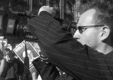 A person with dark hair and glasses, holding a video camera, documenting an anti-war demonstration, holding a video camera surrounded by placards.