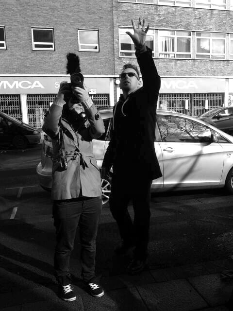 A person with dark hair, glasses, and clothing gesturing upwards as another person films with a video camera and microphone attached, in a street.