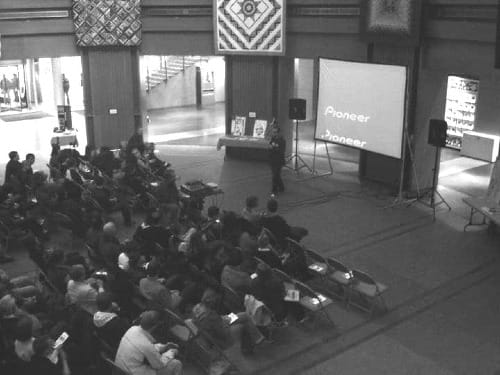 A person in front of a screen, holding a microphone and speaking to a seated audience in an atrium.