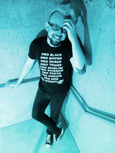 A person with dark glasses, facial hair, and nail polish stood smiling awkwardly at the camera.